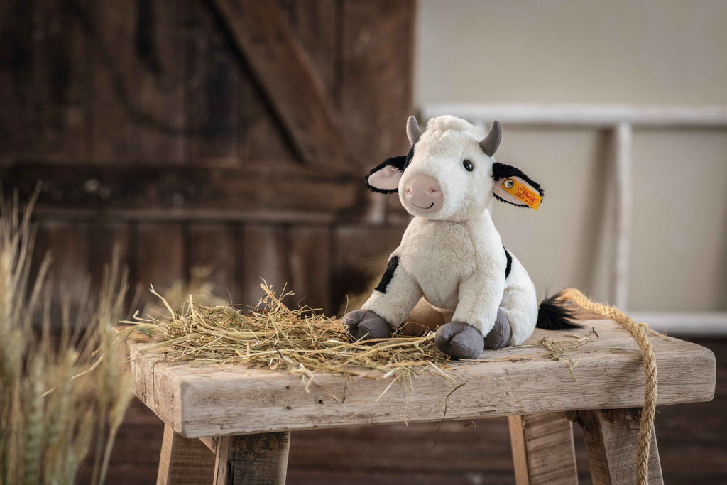 A Steiff cow stuffed animal sitting on a small wooden stool covered with hay, set against a rustic barn background.