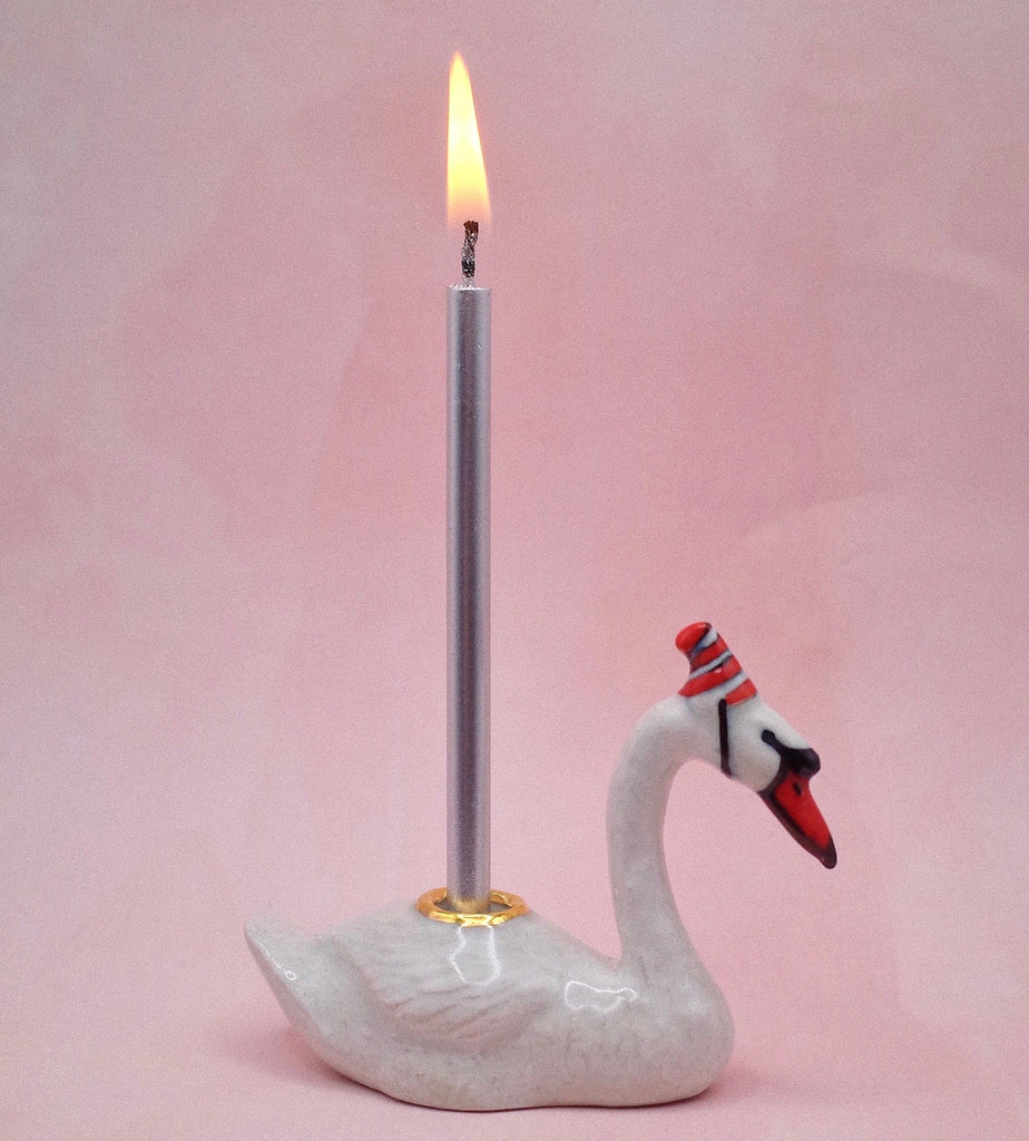 A lit silver candle stands in a Swan Cake Topper candle holder, set against a light pink background.