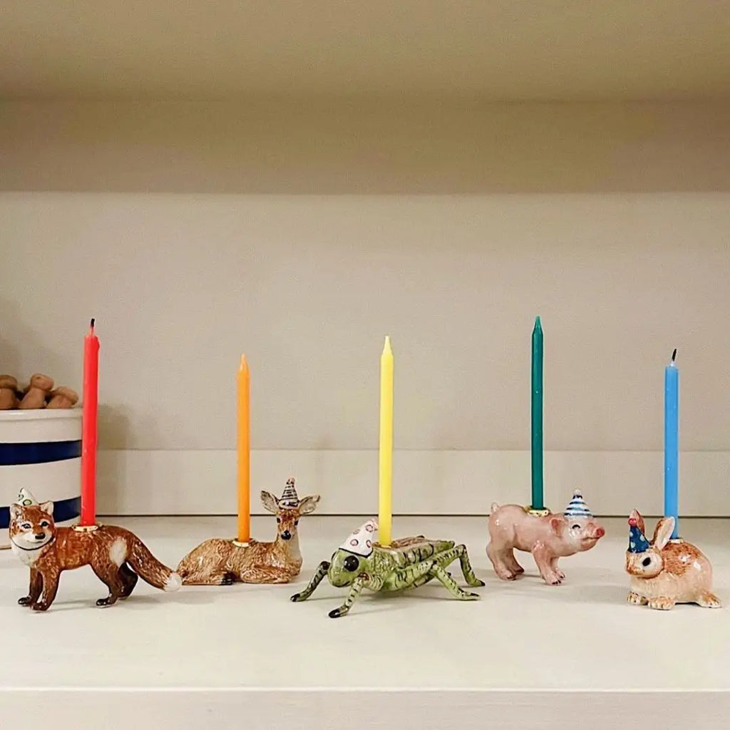A whimsical display featuring the Cricket Cake Topper, a hand-painted porcelain animal figurine (a fox, two deer, and a frog) each wearing a small party hat and balancing colorful birthday candles on their backs, arranged on
