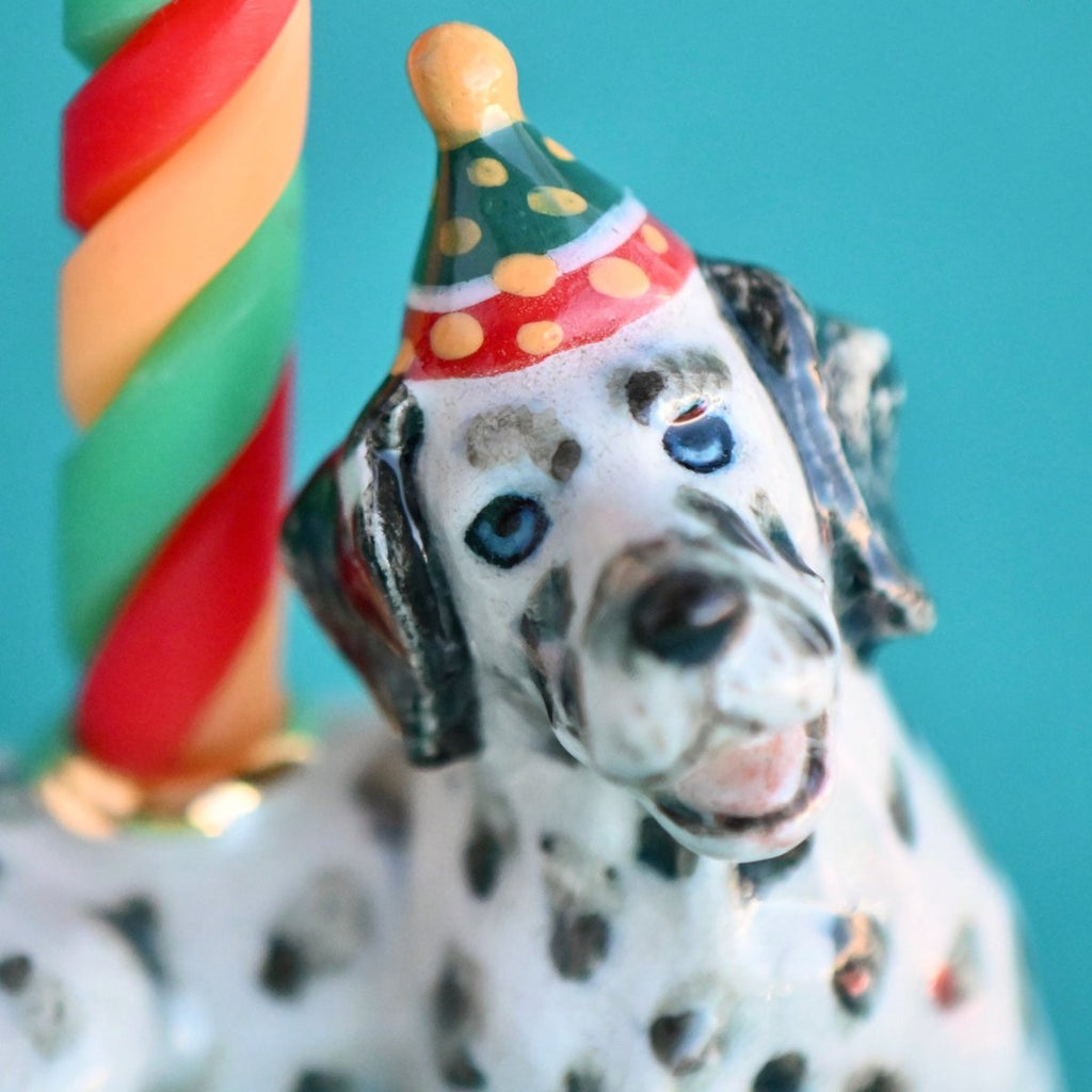 Close-up of a hand-painted Dalmatian Cake Topper with a colorful party hat next to a twisted candle, set against a turquoise background.