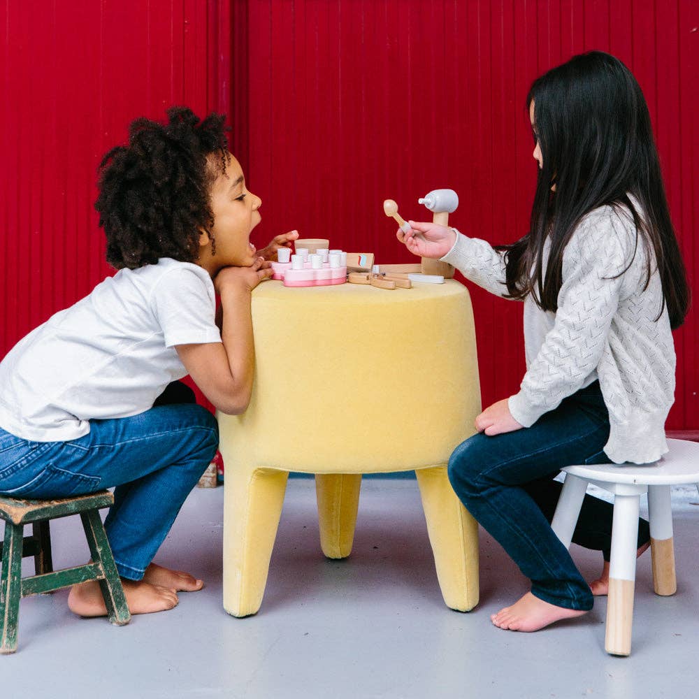 Two children sit on small stools at a round yellow table with a Wooden Dentist Set, pretending to examine teeth and role-play as dentist and patient, against a red background. One child mimics eating or drinking during their imaginative play.