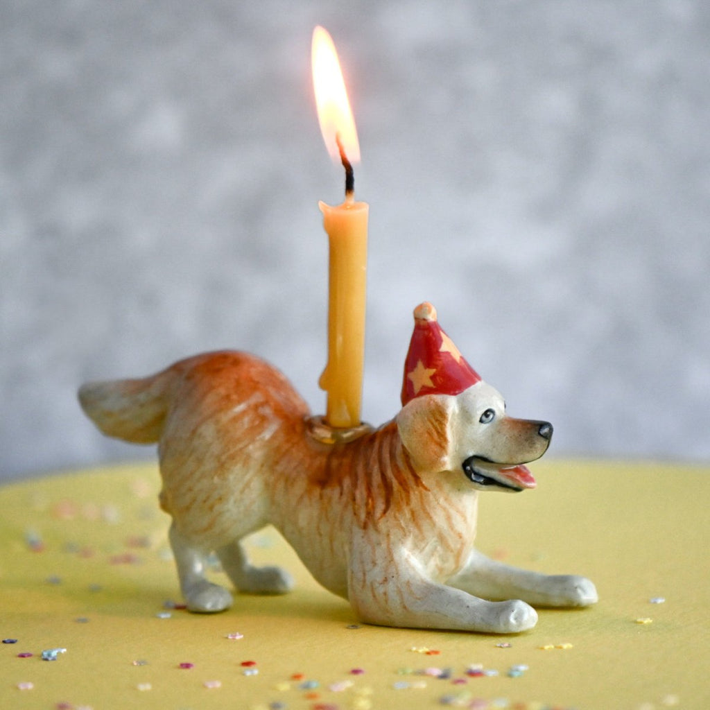 A Golden Retriever Cake Topper of a smiling dog wearing a party hat, with a lit candle mounted on its back, set against a yellow surface with scattered colorful confetti.