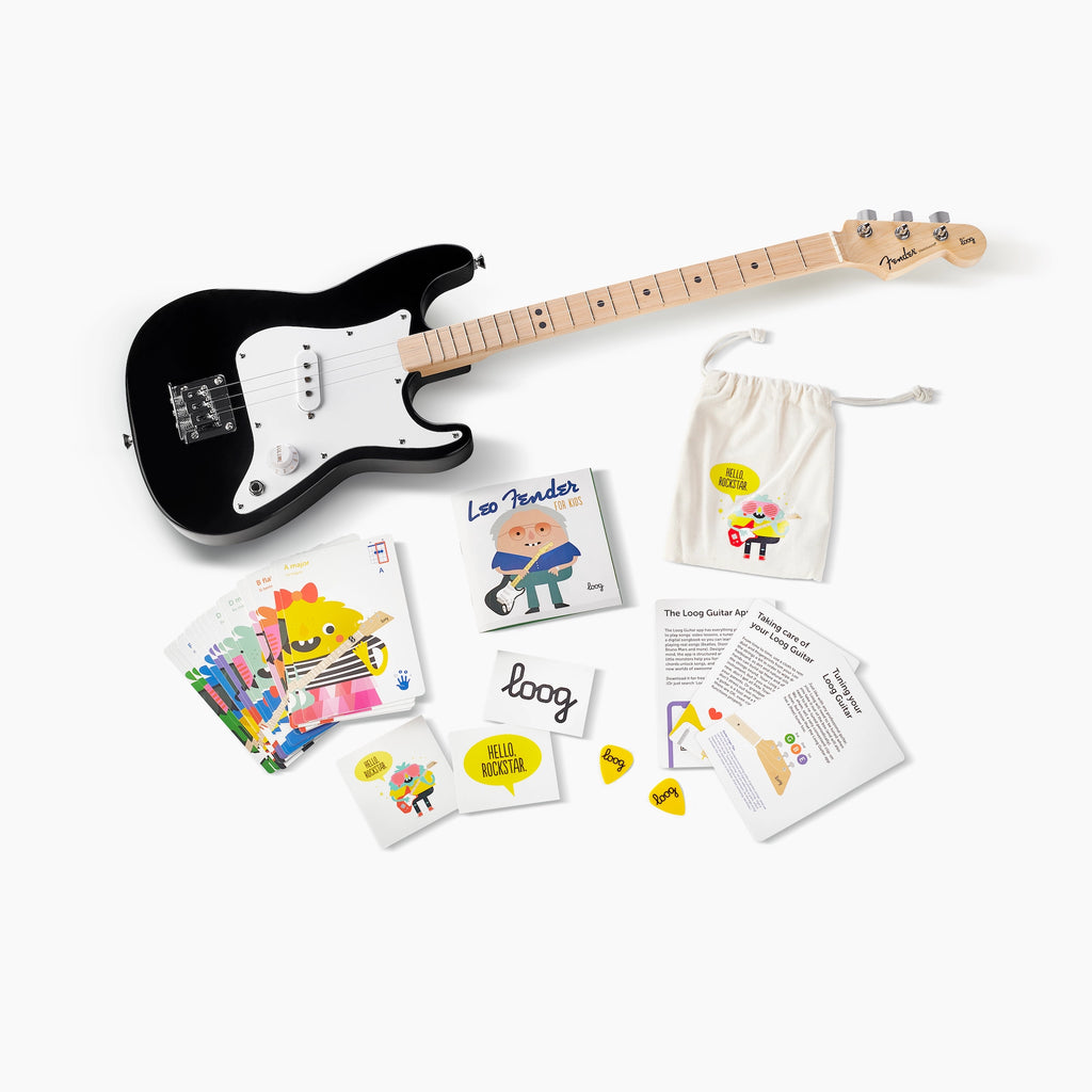 A black and white Fender X Loog Stratocaster Electric Guitar with a maple neck alongside educational materials including flashcards, an illustrated book, and a button pack, all spread out on a plain white background.