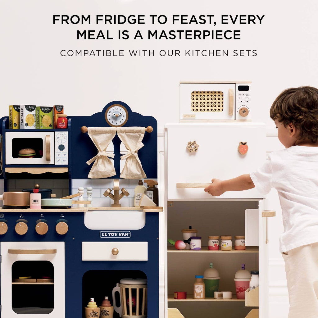 A young child plays with the Wooden Microwave, a sustainable wood play kitchen set. The navy oven, stove, white refrigerator, and accessories inspire imagination. Text above: "From fridge to feast, every meal is a masterpiece." Ships in one week.