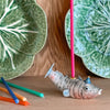 A hand-painted porcelain Fish Cake Topper on a wooden surface, surrounded by colored pencils, with green leaf-patterned plates in the background.