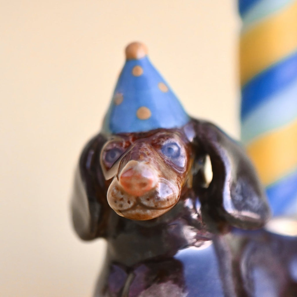 A close-up of a whimsical Dachshund Cake Topper figurine of a dog wearing a blue pointy hat, with a focus on its detailed, glossy face against a soft, blurred background.