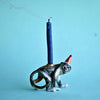 A whimsical Monkey Cake Topper shaped like a monkey wearing a party hat, with a lit beeswax birthday candle on its back against a light blue background. Smoke wisps from the candle's extingu