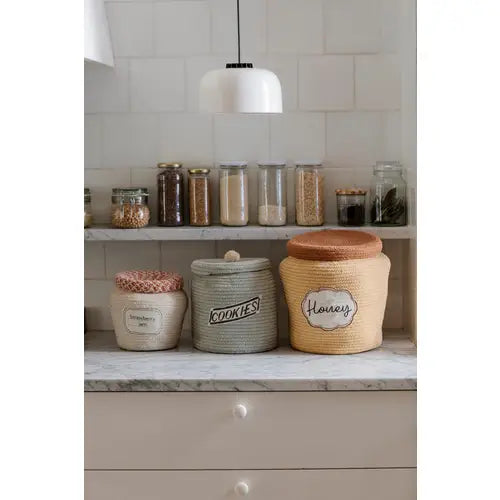 A neatly organized kitchen shelf displaying various labeled containers and jars with ingredients like cookies, honey, and Strawberry Jam Jar Basket, against a tiled backsplash under a hanging white lamp.