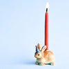 A hand-painted ceramic Rabbit/Bunny Cake Topper with a party hat holding a lit red taper candle, against a light blue background.