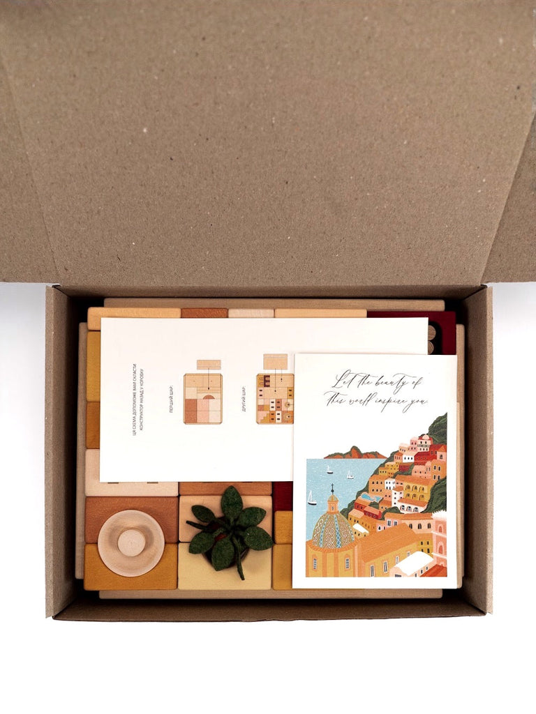 An open cardboard box containing an assortment of items including colorful Sabo Concept Italian Courtyard Blocks, postcards with artistic designs, and a small green plant, arranged neatly on a white background.