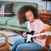A young girl with curly hair smiles joyfully while playing a Fender X Loog Stratocaster Electric Guitar, sitting on a stool in a cozy living room.