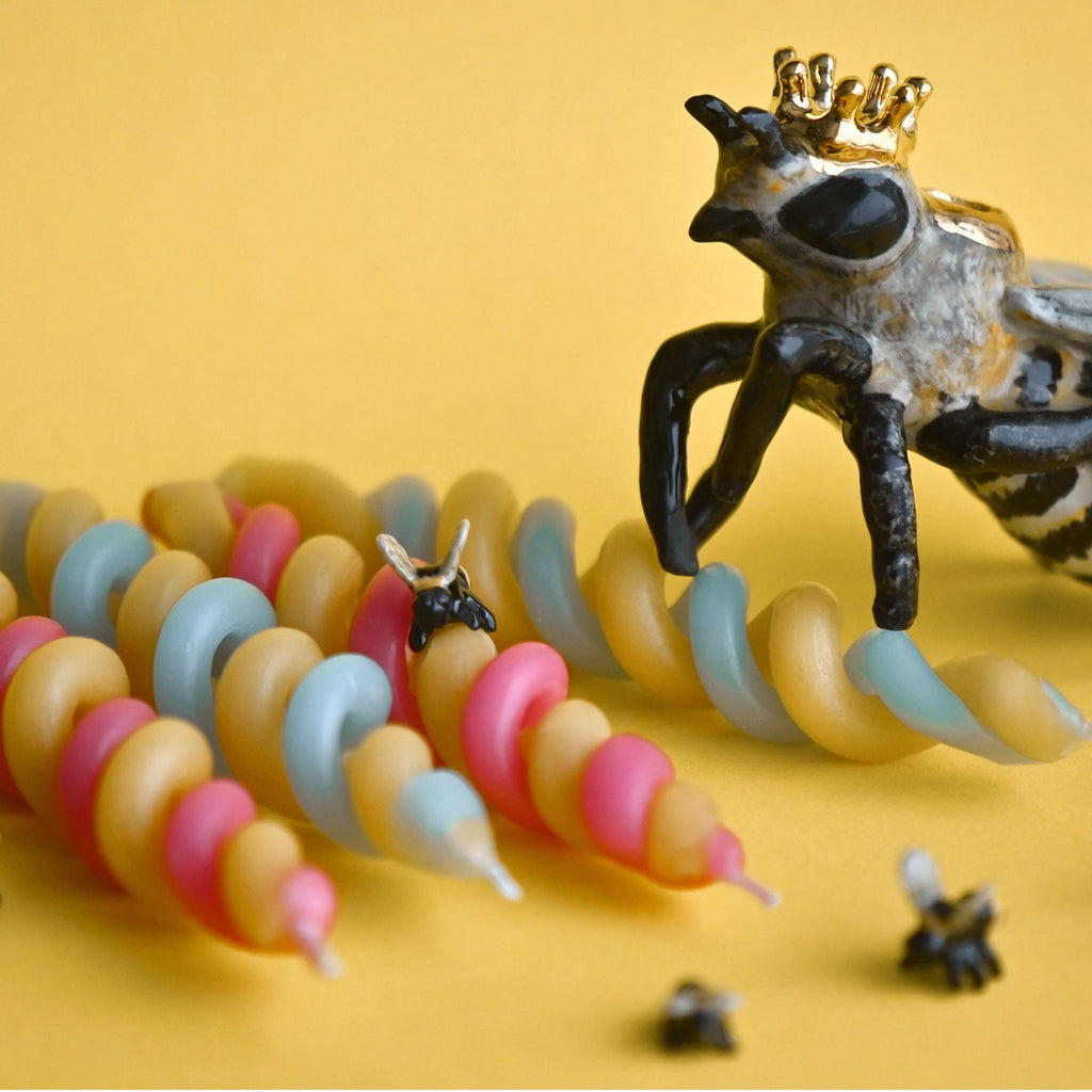 A whimsical fine porcelain Queen Bee Cake Topper with a crown, surrounded by colorful pastel beads and miniature metal crowns on a yellow background.