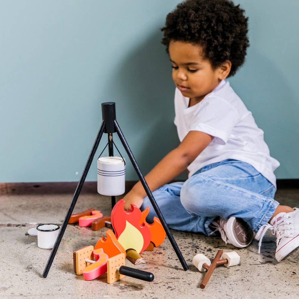 A young child sits on the floor playing with the Wooden Camp Fire Set, which includes a camp fire, pot, utensils, and food items under a tripod—ideal for indoor camping and imaginative pretend play.