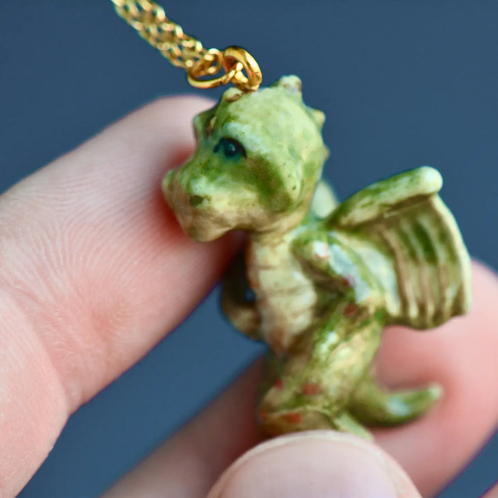 A close-up view of a small handcrafted porcelain Baby Dragon Necklace being held between fingers, showcasing detailed green and gold textures on a dark background.