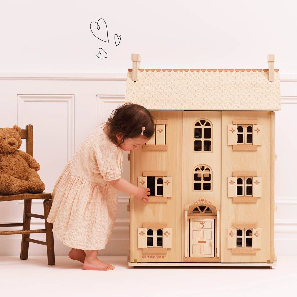 A young child in a light dress plays imaginatively with the windows of The Grand Natural Wood Doll House, while a brown teddy bear sits on a chair nearby beneath two heart drawings on a white wall.