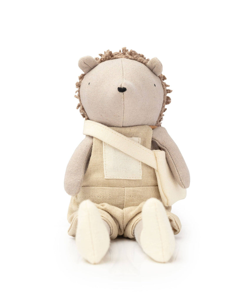 Heather The Hedgehog plush toy features a beige fabric body and brown yarn spikes, wearing tan overalls with a white patch. A small beige bag is slung over her shoulder. It sits upright, exuding enchanting charm against a plain white background, ready for garden escapades.