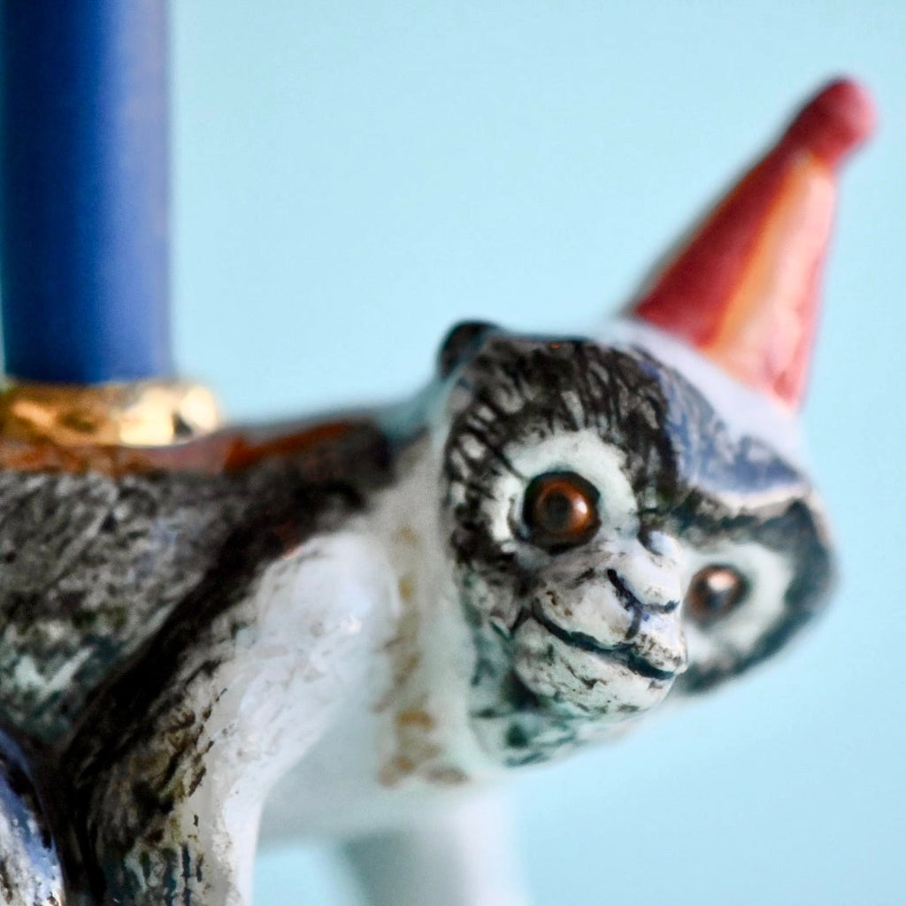 A close-up image of a hand-painted Monkey Cake Topper, painted in shades of white, grey, and black with gold accents, wearing a red pointed hat.