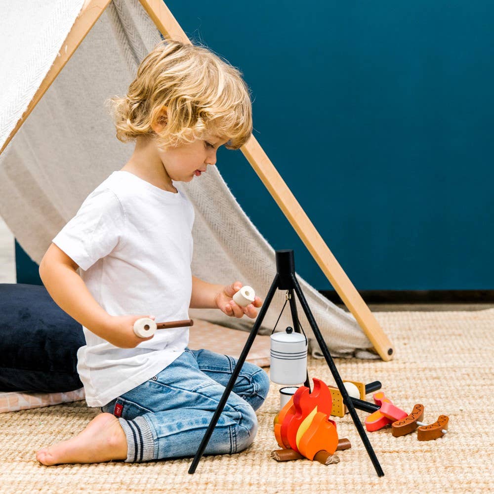A young child enjoys indoor camping with the Wooden Camp Fire Set, holding toy marshmallows over play flames, wooden logs, and a hanging pot beside a tent on a woven rug.