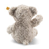A Steiff, Honey Teddy Bear, 11 Inches with curly fur, a button in the ear, looking upwards and waving with its left paw, isolated on a white background.