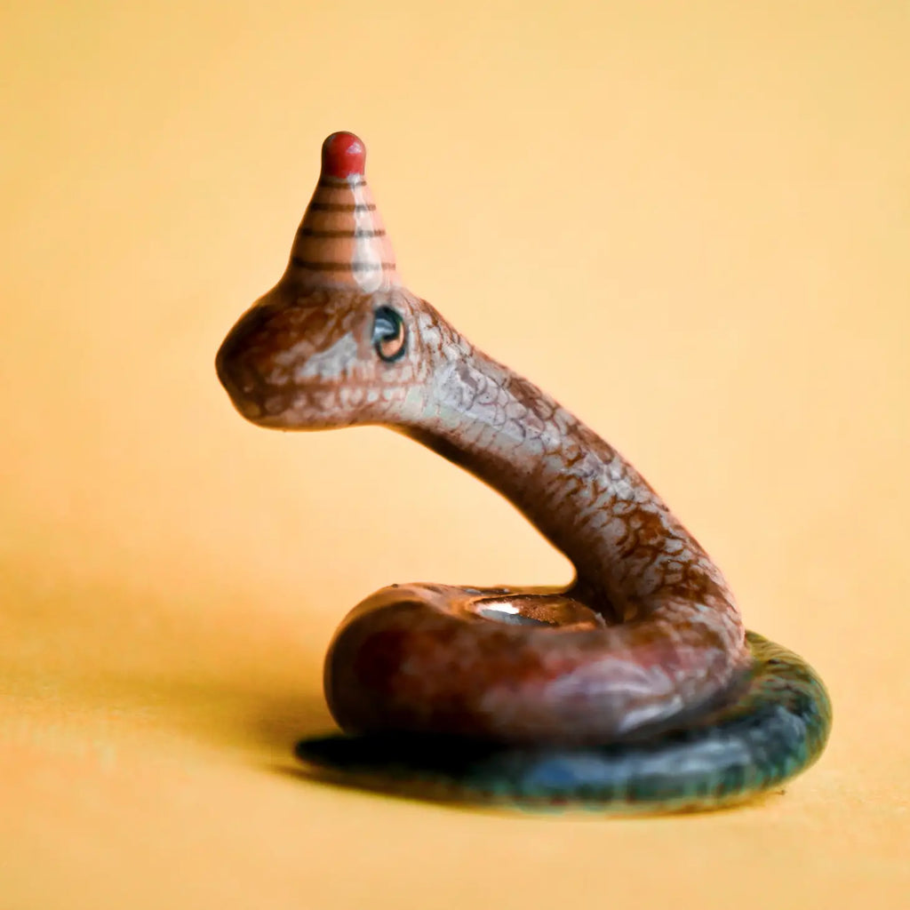 A close-up photograph of a small, intricately detailed hand-painted Snake Cake Topper figurine of a brown snake with red and blue accents, coiled on a soft yellow background.