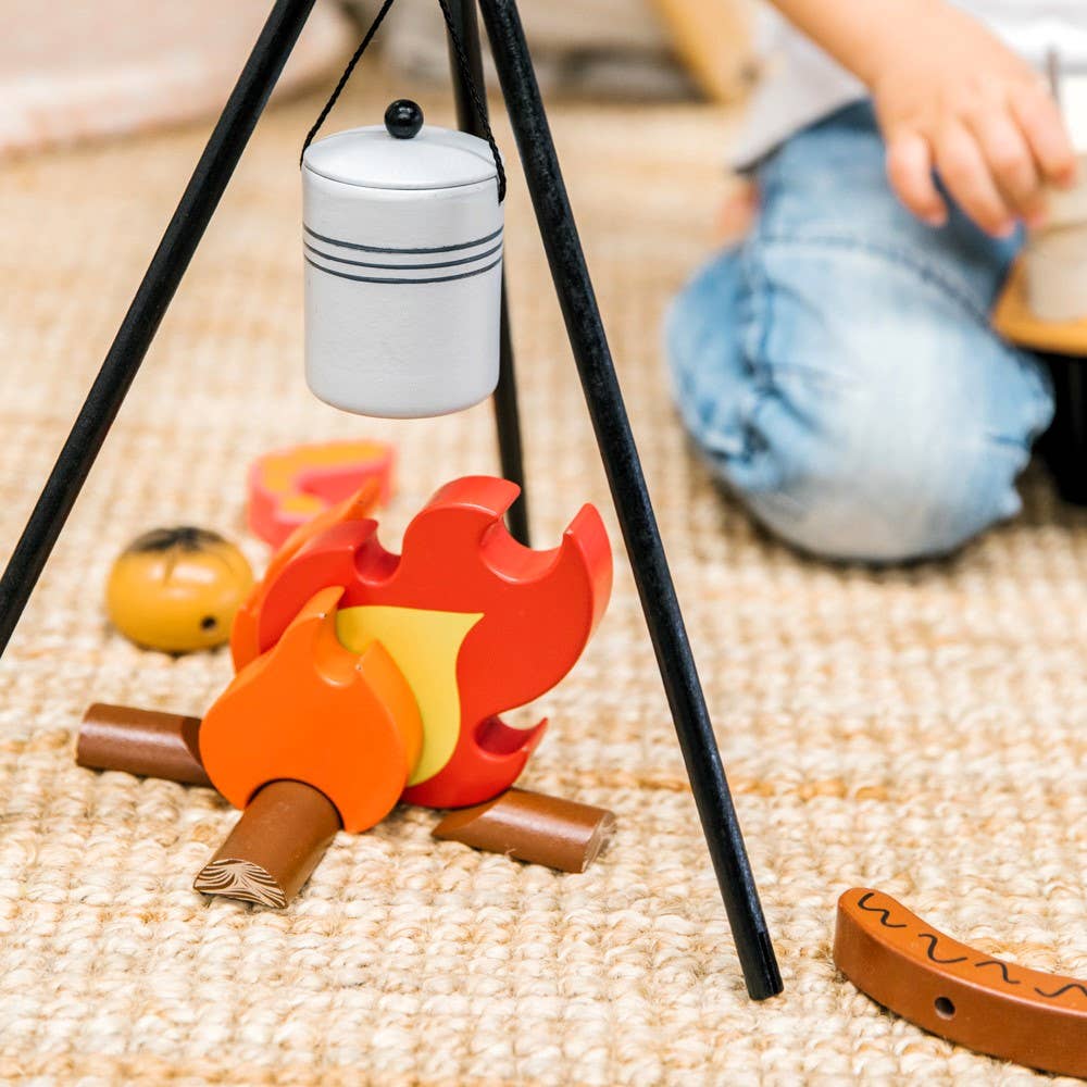 A child sits on a woven rug beside the Wooden Camp Fire Set, which includes wooden flames, logs, and a pretend pot—perfect for indoor camping fun and imaginative kids' pretend play.