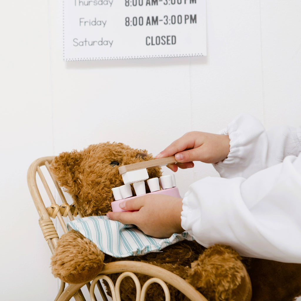 A child in a white shirt pretends to brush a brown teddy bear’s teeth using the Wooden Dentist Set. The teddy sits in a small chair with a striped cloth, and a business hours sign appears in the background.