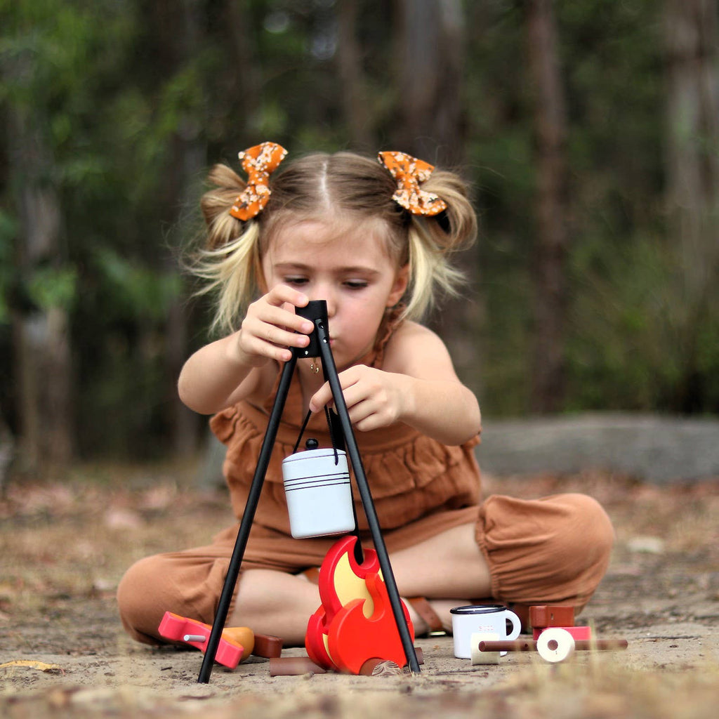 A young child with pigtails and orange bows sits outdoors, enjoying the Wooden Camp Fire Set for kids’ pretend play, featuring a toy fire, mugs, and a pot over the fire amid trees and nature.