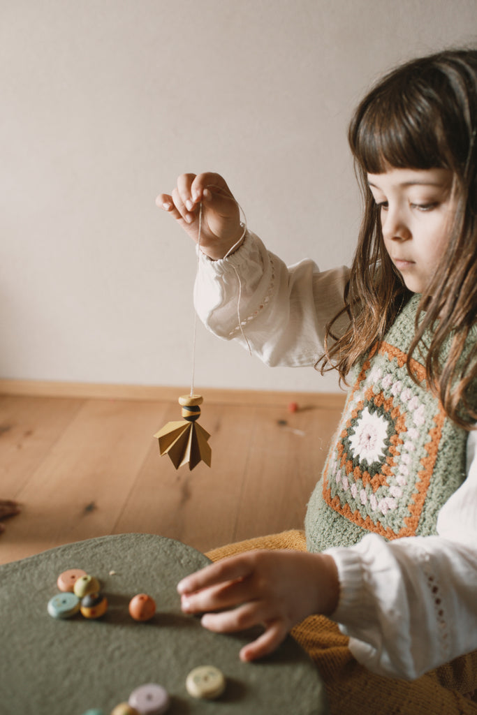 A young child with long brown hair holds up a wooden spinning top by a string, sitting at a table with colorful pieces from the Grapat Quiet Time Advent Calendar 2025, wearing a green crocheted vest over a white shirt.