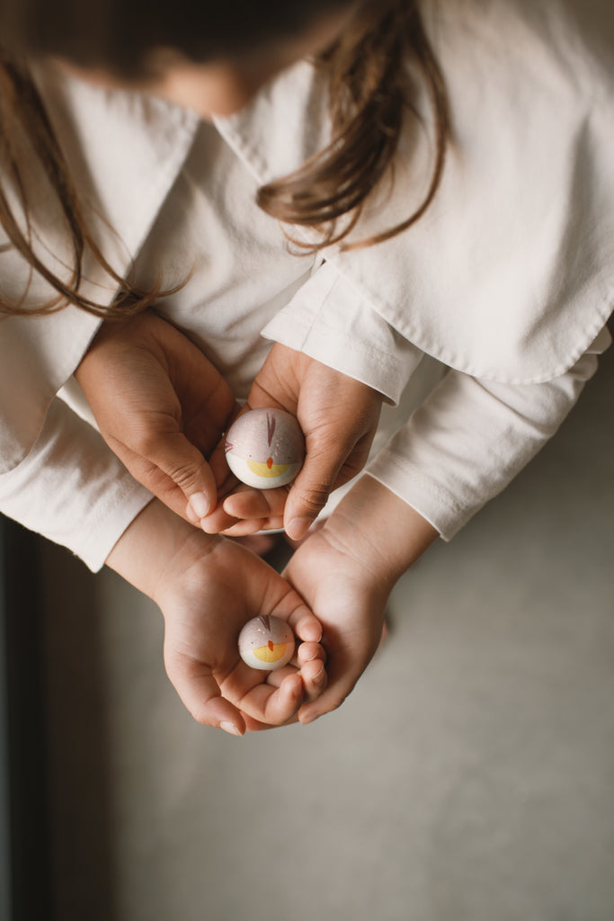 An adult's hands gently hold a child's as they cradle a small, chick-painted egg from the limited-edition Grapat Quiet Time Advent Calendar 2025. Both wear white, with the focus on their hands and the colorful egg.