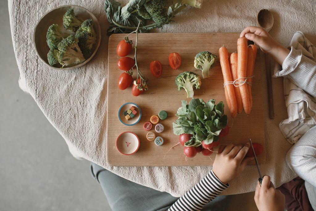 Two people share peaceful kitchen moments with the Grapat Quiet Time Advent Calendar 2025, preparing fresh broccoli, tomatoes, radishes, and carrots on a wooden cutting board over a beige cloth, fully embracing quiet time together.