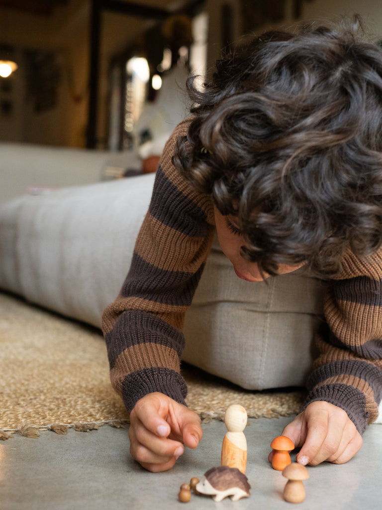 A young child with curly hair lies on the floor, playing with small wooden toys from the Hello Autumn Set (Grapat X Ostheimer Limited Edition), including a turtle, mushroom, and peg dolls, in a cozy living room.