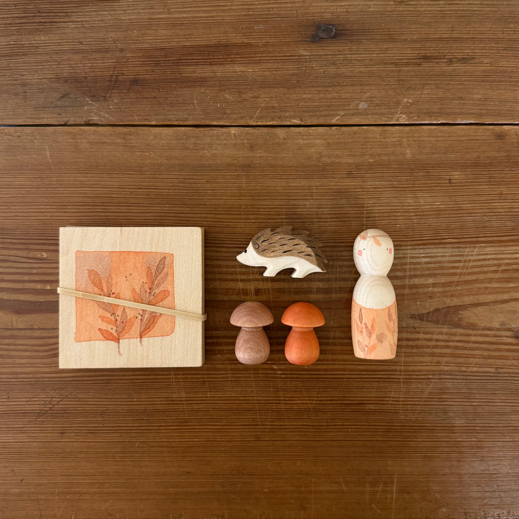 The Hello Autumn Set (Grapat X Ostheimer Limited Edition) features a square block with leaf design, elastic band, small hedgehog, two mushroom-shaped pieces, and a painted peg doll with orange leaf pattern on a wooden surface.
