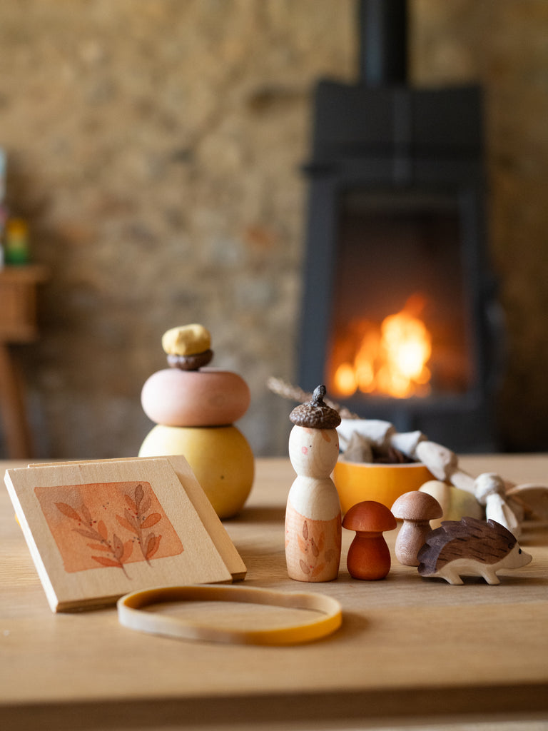The Hello Autumn Set (Grapat X Ostheimer Limited Edition) features wooden autumn-themed toys—a doll with an acorn cap, mushrooms, a hedgehog, and a framed leaf—arranged on a table by a lit fireplace against a stone wall.