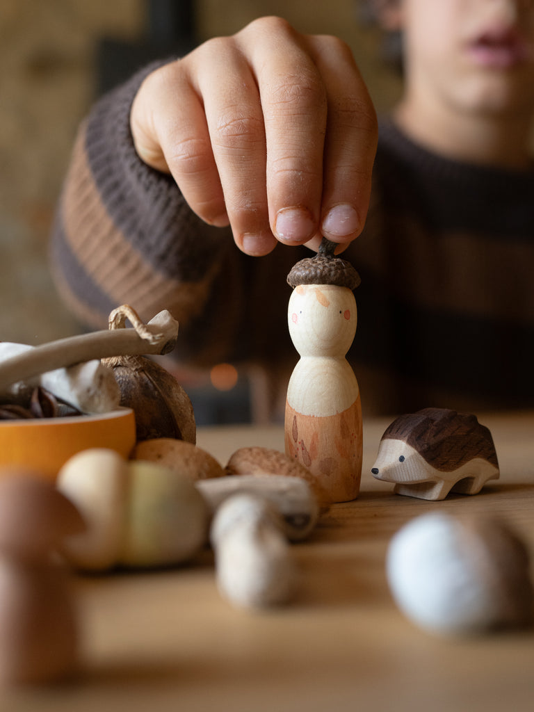 A child in a brown striped sweater places an acorn cap on a small figure from the Hello Autumn Set (Grapat X Ostheimer Limited Edition), surrounded by autumn-themed toys, nuts, and a wooden hedgehog on the table.