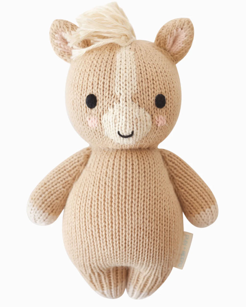 The Cuddle + Kind Baby Pony is a hand-knit beige plush toy made from soft Peruvian cotton yarn, with cream accents, black stitched eyes, a smiling face, blush cheeks, a yarn mane, and cuddly round arms and legs.
