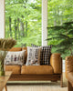A cozy living room scene with a stylish tan leather sofa adorned with various Trick or Treat Pillows. Large windows display a lush green forest backdrop.
