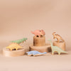 A collection of Handmade Wooden T-rex Dinosaur toys displayed on circular wood platforms with a beige background. The dinosaurs are painted in various pastel colors using non-toxic paint.