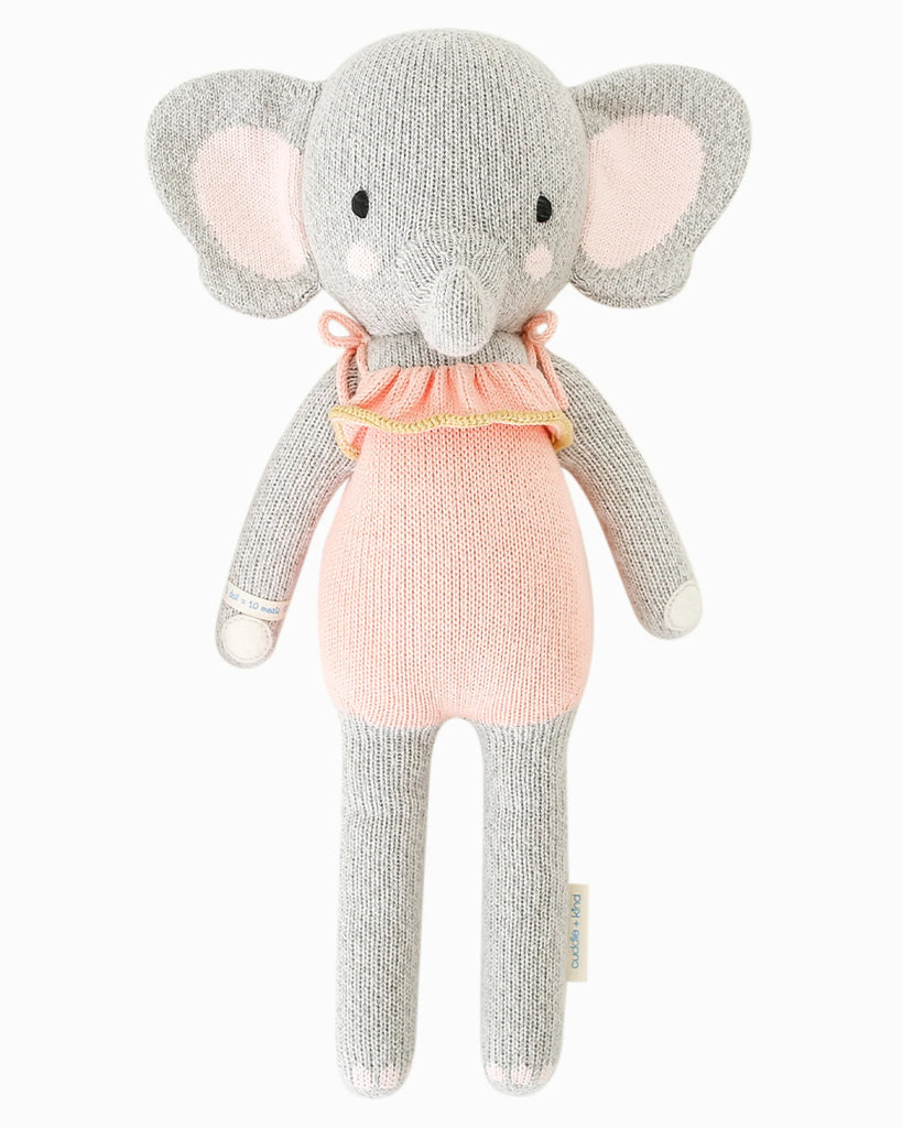 Cuddle + Kind Eloise the Elephant is a hand-knit doll featuring soft gray and light pink colors, large ears, a pink dress, stitched details, and a small side tag—capturing charming savanna style in an adorable stuffed animal.