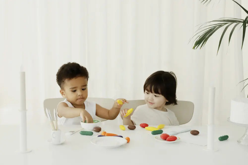 Two young children are sitting at a white table, engaging in play with Farm Crayons and toys. The setting has a minimalist aesthetic with white curtains in the background.