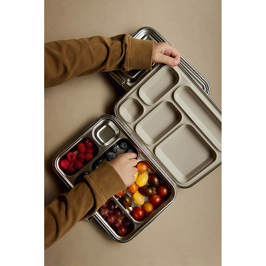A person wearing a brown long-sleeve shirt arranges various fruits, including raspberries, blueberries, grapes, and cherry tomatoes, into the compartments of a Haps Nordic - Stainless Steel Lunch Box - Multi-Compartment (ships in approximately two weeks) for meal prep.