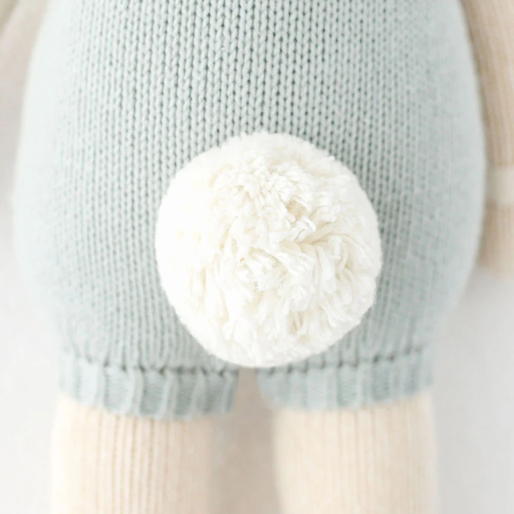Close-up of the back of a Cuddle + Kind Henry the Bunny hand knit doll, made from 100% cotton yarn with light blue fabric and a large, fluffy white yarn pom-pom tail.