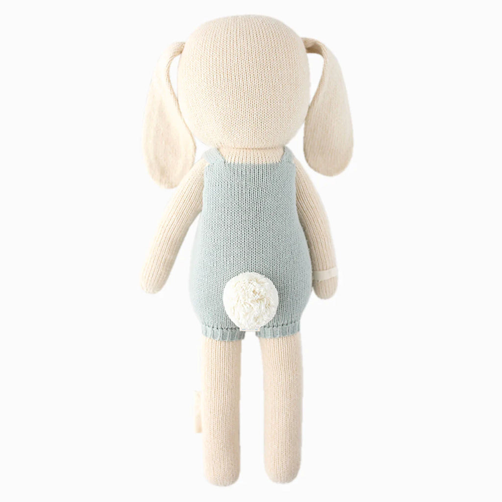 The Cuddle + Kind Henry the Bunny is a hand-knit plush bunny made from 100% cotton yarn, featuring floppy ears, a light blue outfit, and a small white tail, shown from the back against a plain white background.