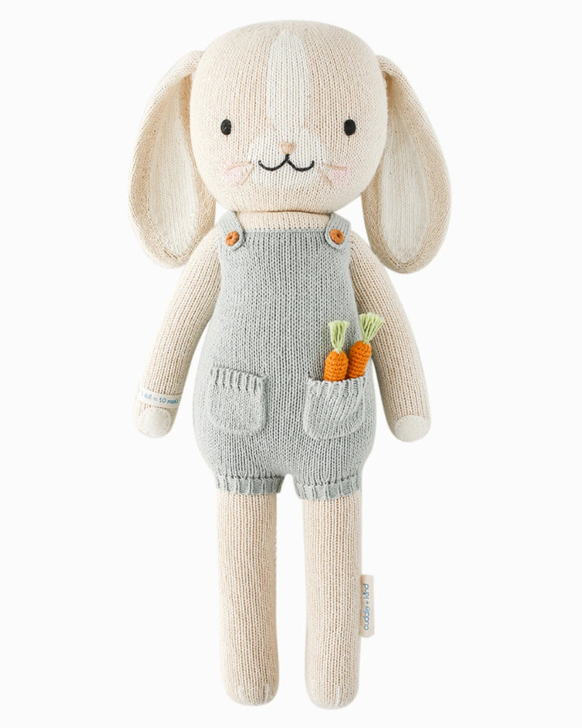 Cuddle + Kind’s Henry the Bunny is a hand-knit, fair trade stuffed animal with cream fur, long ears, and a smiling face. He wears blue overalls with two small carrots in his pocket for extra charm.