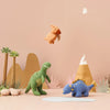 A playful display featuring Olli Ella Holdie Pre-Historic Animals on a sandy backdrop with small paper plants, a toy volcano, and a plush bear seemingly leaping in the air. Soft.
