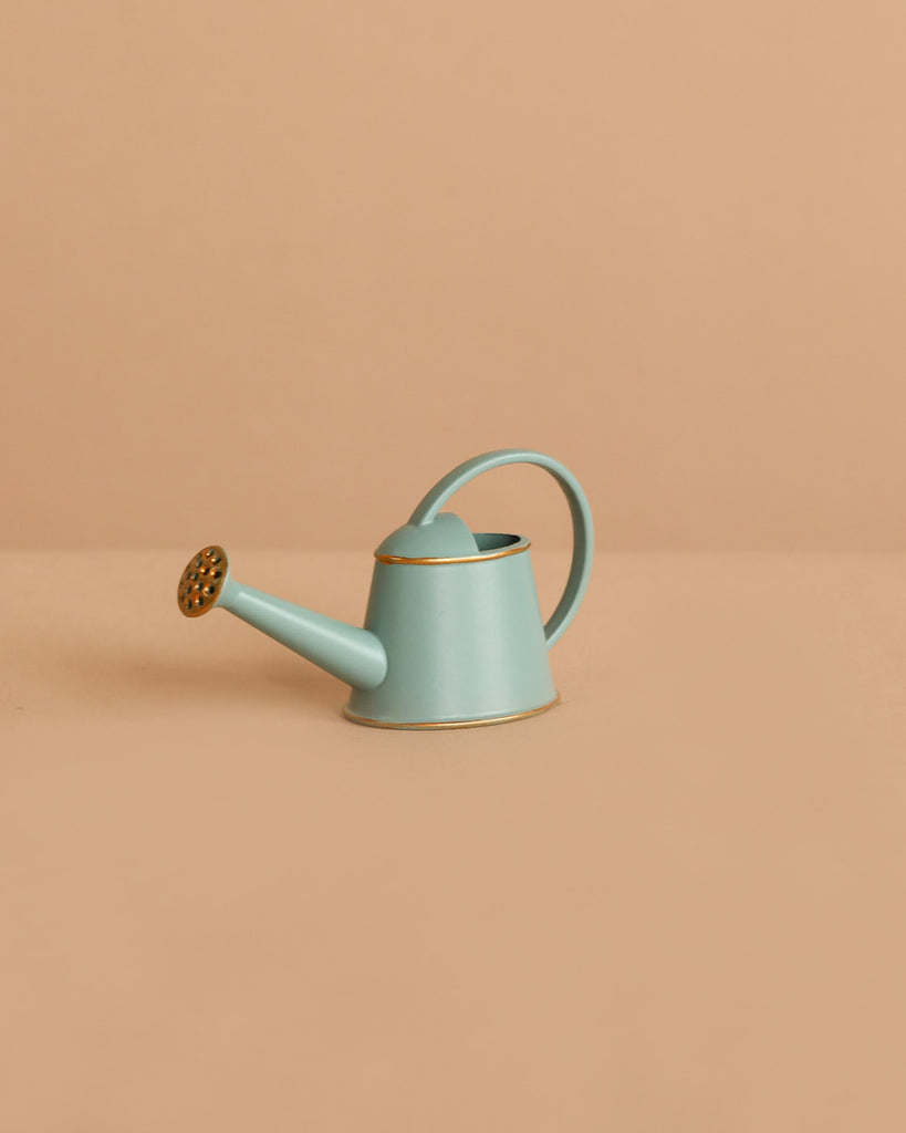 The Maileg Watering Can, Mouse - Light Blue, is a small metal accessory with a gold spout and trim, displayed on a beige surface and background—ideal for adding charm to any play scene.