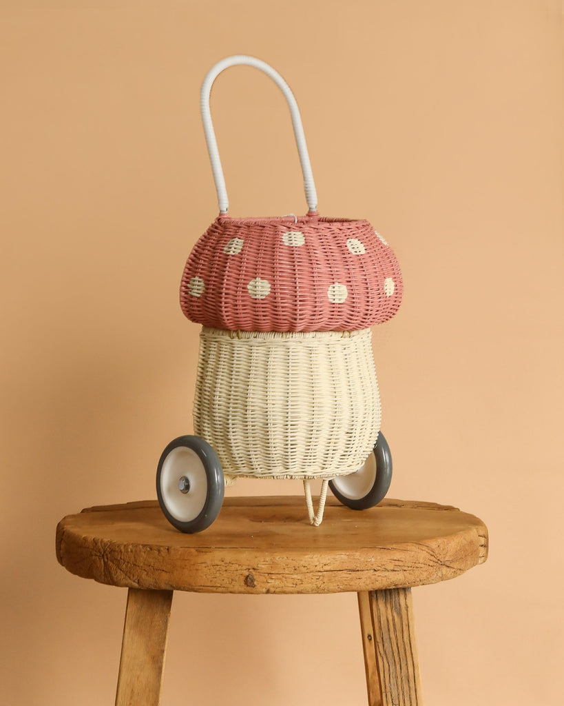 The Olli Ella Rattan Mushroom Luggy - Pink, shaped like a mushroom with a pink and white polka-dot top and cream base, sits on a wooden stool—ideal for a woodland-themed nursery. Features include a white handle, two wheels, and light brown accents.