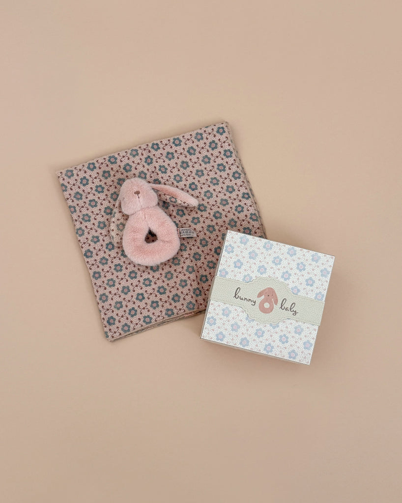 The Maileg Bunny Rattle Set - Powder features a small pink stuffed bunny on a pink floral blanket in a charming gift set. Next to it is a box labeled "bunny baby" with blue flowers, all set against a soft beige background.