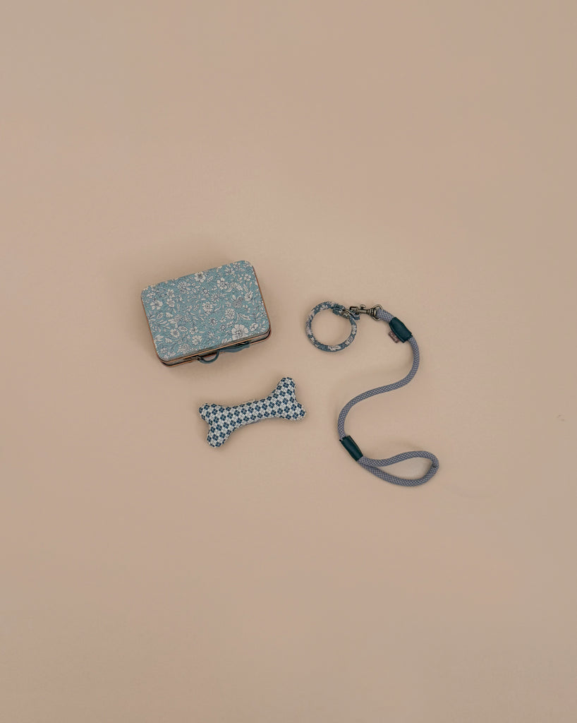 The Maileg Puppy Supply Accessories - Blue, including a metal suitcase, blue dog bone toy with a geometric pattern, and matching light blue leash and collar, are arranged on a beige background.