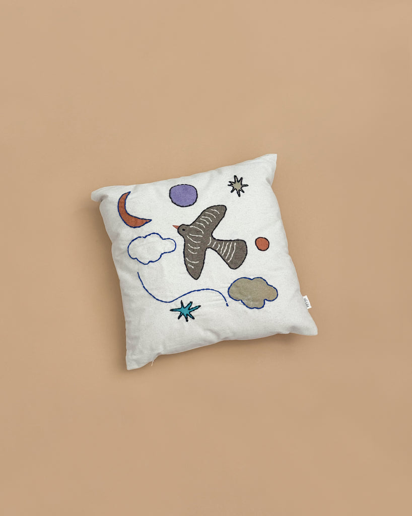 The Ferm Living Naïve Cushion - Bird is a square kids cushion made of light organic cotton, featuring hand-embroidered designs of a bird, clouds, stars, a crescent moon, and circles on a plain beige background.