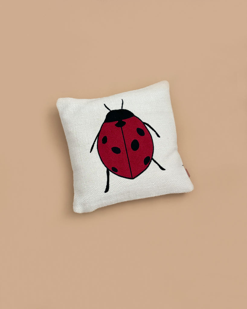 The Ferm Living Forest Embroidered Cushion - Ladybird is a white organic cotton cushion with a simple red ladybug featuring black spots and legs, set on a beige background.